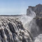 A stunning view of Dettifoss waterfall in Iceland, showcasing its powerful cascade as it plunges dramatically into a rugged canyon. The waterfall, known as Europe's most powerful, creates a misty spray that catches the sunlight, forming a rainbow in the air. Surrounding the waterfall, steep cliffs and rocky terrain highlight the natural beauty and raw power of this iconic landmark, while the lush greenery in the distance adds a touch of color to the scene.