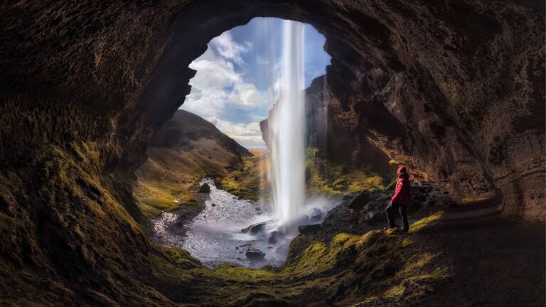 A man stands behind Kvernufoss waterfall, surrounded by a misty curtain of water cascading down a rocky cliff. The vibrant green moss covers the surrounding rocks, creating a lush backdrop, while the sunlight filters through, casting shimmering reflections on the water. The man, silhouetted against the waterfall, gazes out at the serene landscape, embodying a sense of adventure and connection to nature in this breathtaking setting.