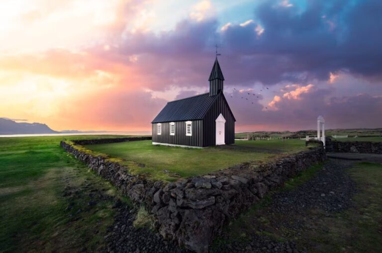 A stunning sunset view of Búðarkirkja, the iconic black church in Búðir, Snæfellsnes Peninsula, Iceland. The church stands boldly against a vibrant sky painted with hues of orange, pink, and purple, while the silhouette of the surrounding mountains adds depth to the scene. The church's dark exterior contrasts beautifully with the warm colors of the sunset, creating a serene and picturesque atmosphere that highlights the tranquil beauty of this historic site. Lush green fields surround the church, completing this enchanting landscape.