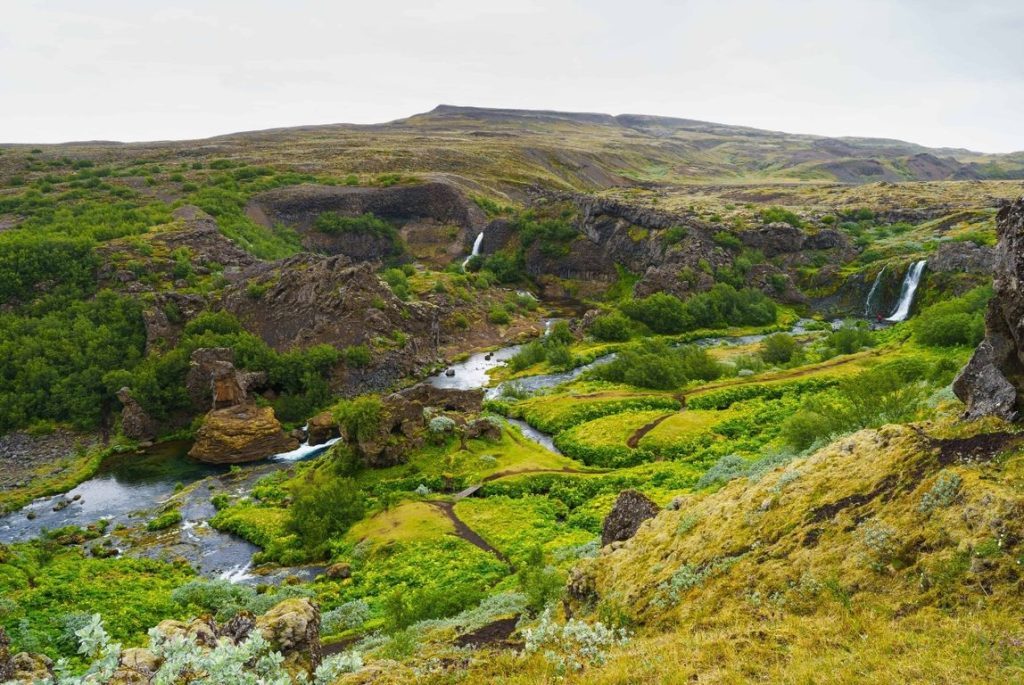 Aerial View of Gjáin: An aerial perspective of Gjáin, revealing the lush oasis nestled within a rugged volcanic landscape. The photo captures the winding stream, multiple waterfalls, and vibrant greenery that define the valley. Surrounded by jagged cliffs and barren terrain, Gjáin appears as a hidden, fertile gem in the midst of Iceland’s wild nature.