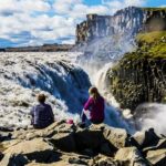 A man stands in awe at the edge of Dettifoss waterfall in Iceland, gazing at the thundering cascade as it crashes into the rocky gorge below. The powerful flow of water creates a mist that envelops the area, while rainbows form in the spray under the sunlight. The rugged landscape, with its steep cliffs and boulders, frames the scene, showcasing the natural beauty and intensity of this iconic waterfall.