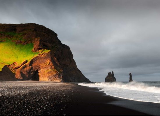 A breathtaking view of Reynisfjara black sand beach in Iceland, showcasing the striking Reynisdrangar cliffs in the background. The dark volcanic sand stretches across the foreground, while powerful waves crash against the shore. The towering, jagged cliffs rise majestically from the ocean, contrasting beautifully with the deep blue water and cloudy sky. The scene captures the wild and rugged beauty of Iceland's coastline.